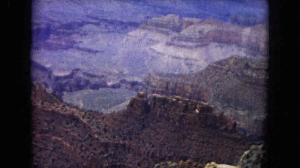 1967 Arizona 8mm frame showing layered Grand Canyon walls, distant mesas, rocky foreground, and a hazy blue-purple sky.
