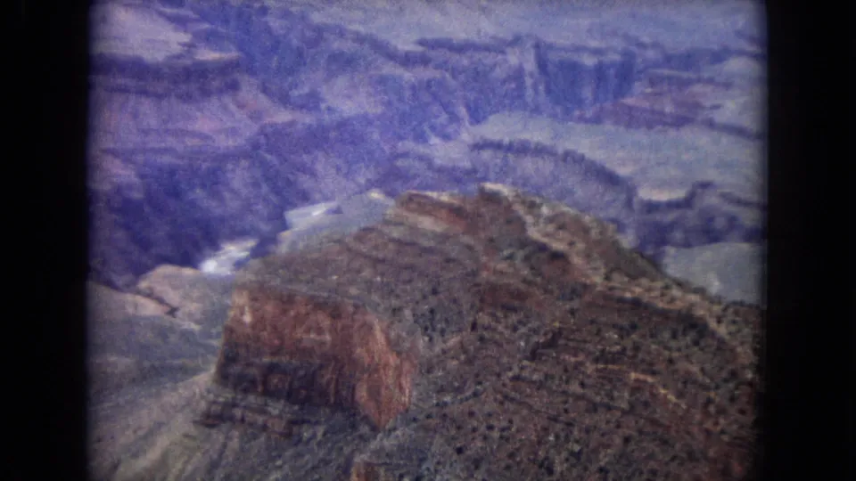 1967 Arizona home-movie frame showing a reddish canyon butte, sloped desert terrain, and layered Grand Canyon walls beyond.