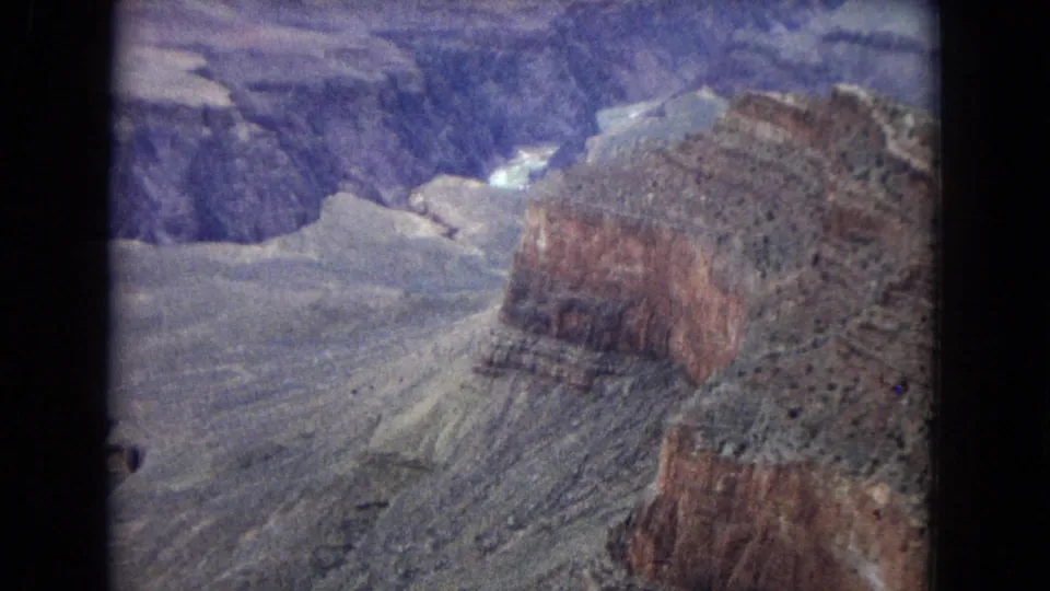 1967 Arizona 8mm frame of a large stratified Grand Canyon formation, pale desert slopes, and deep blue-purple canyon recesses.
