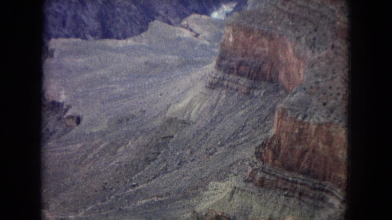 1967 Arizona home-movie frame showing an aerial view over Grand Canyon terrain with pale rock shelves, reddish cliff bands, and soft 8mm color fading.