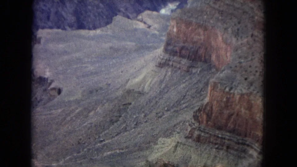 1967 Arizona home-movie frame showing an aerial view over Grand Canyon terrain with pale rock shelves, reddish cliff bands, and soft 8mm color fading.