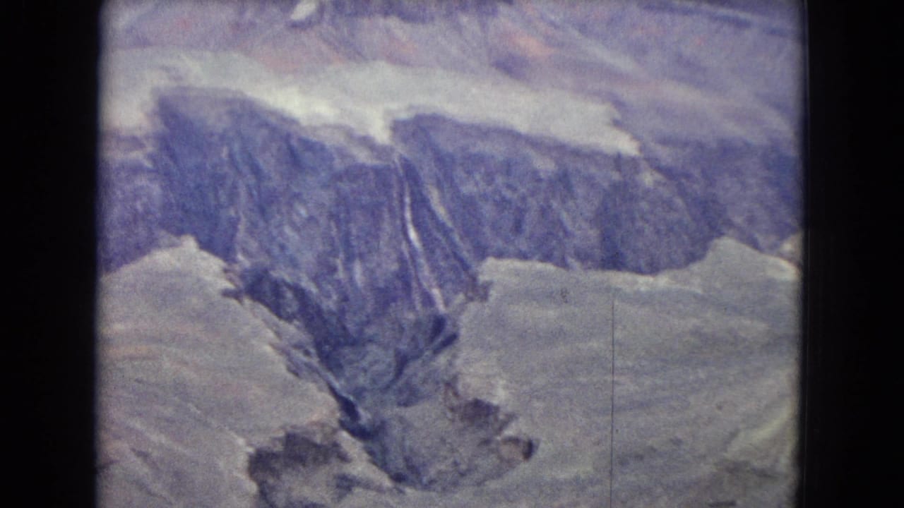1967 Arizona frame showing a high view into the Grand Canyon, with shadowed blue-purple cliff faces, pale rock shelves, and a deep central ravine.
