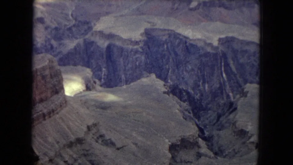 1967 Arizona home-movie frame looking across Grand Canyon cliffs, pale terraces, and a dark ravine, with a bright film or light reflection near the lower left edge.