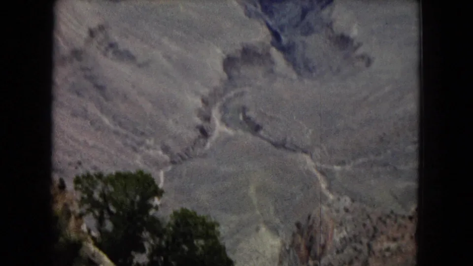 1967 Arizona frame showing pale Grand Canyon terrain from above, with branching ravine lines and dark green trees partly visible along the lower edge of the frame.