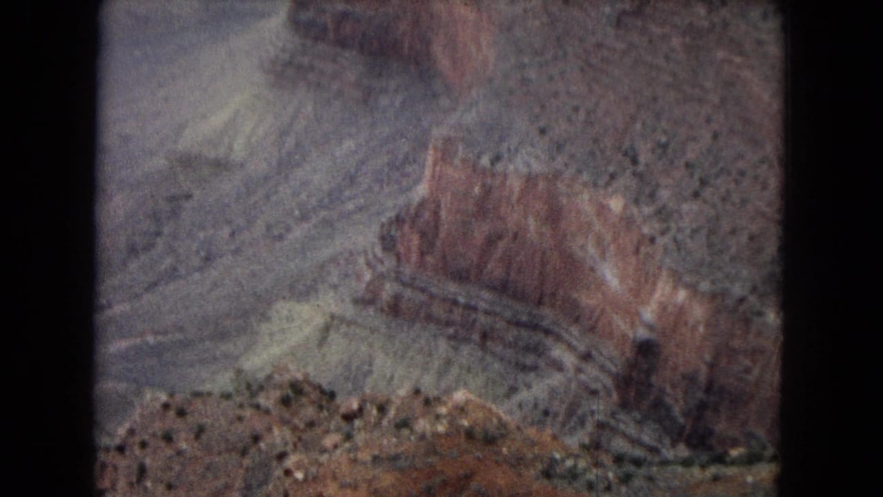 1967 Arizona 8mm travel frame showing red and gray canyon walls, sloping strata, and scattered desert shrubs under soft vintage color.