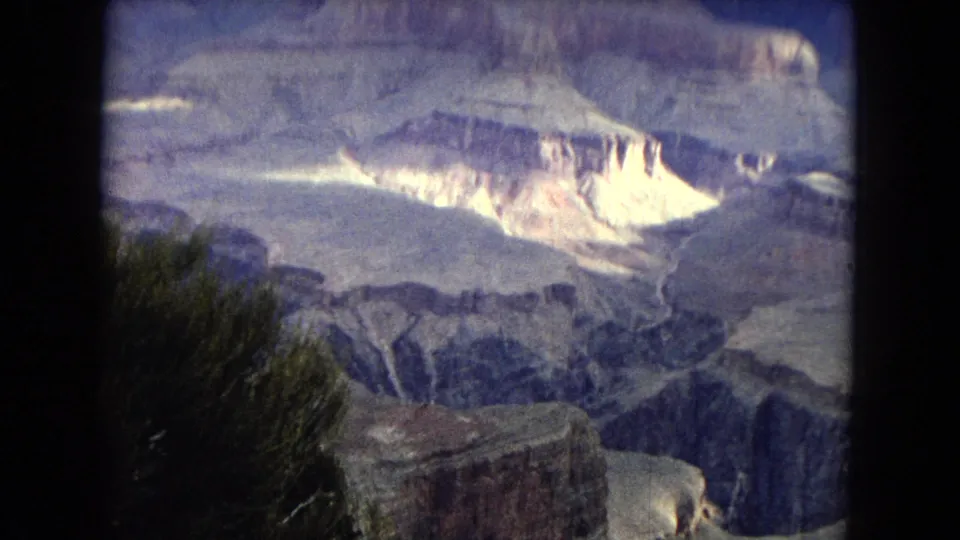 1967 Arizona 8mm frame looking over a wide Grand Canyon landscape with bright sunlit cliffs, deep shadowed ravines, and foreground desert brush.