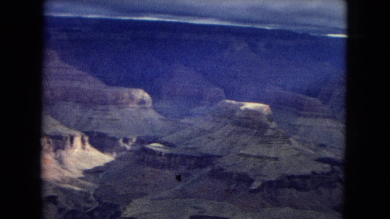 1967 Arizona home-movie frame showing a wide Grand Canyon vista with layered plateaus, pale cliffs, and heavy cloud cover near the horizon.