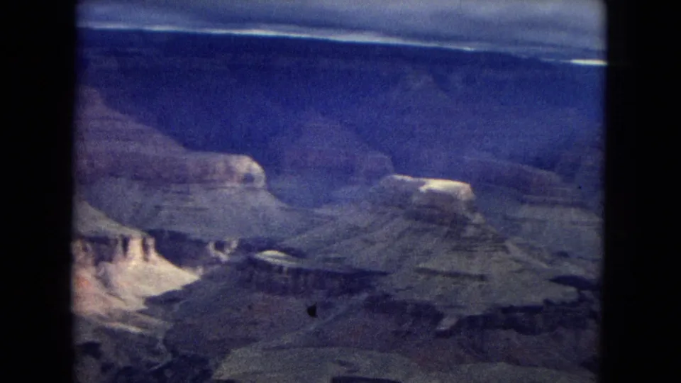 1967 Arizona home-movie frame showing a wide Grand Canyon vista with layered plateaus, pale cliffs, and heavy cloud cover near the horizon.