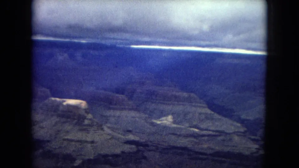 1967 Arizona 8mm frame with a broad canyon plateau, deep valley floor, distant cliffs, and a bright strip of sky beneath darker clouds.