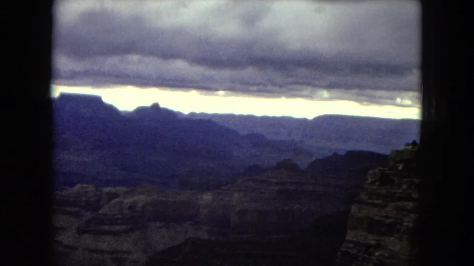 1967 Arizona home-movie frame showing shadowed Grand Canyon ridges, an overcast sky, and a bright horizon break at the end of the pan.
