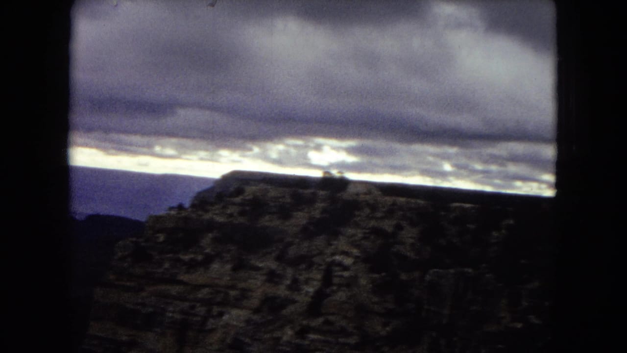 1967 8mm frame of the Grand Canyon with a dark foreground rock formation, distant canyon layers, and a luminous dusk sky.