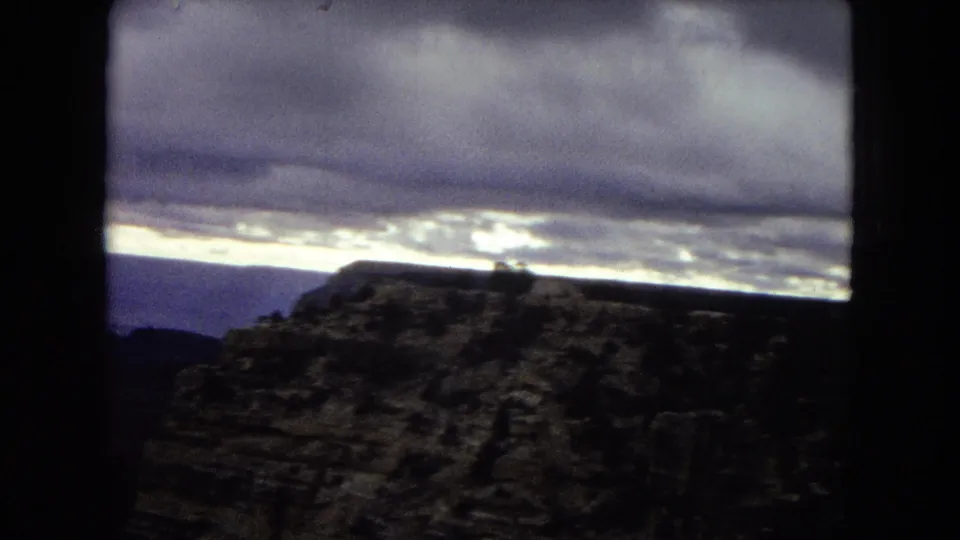 1967 8mm frame of the Grand Canyon with a dark foreground rock formation, distant canyon layers, and a luminous dusk sky.