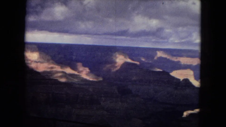 1967 8mm home-movie view across the Grand Canyon, with layered cliffs catching dim light below a dense cloud ceiling.
