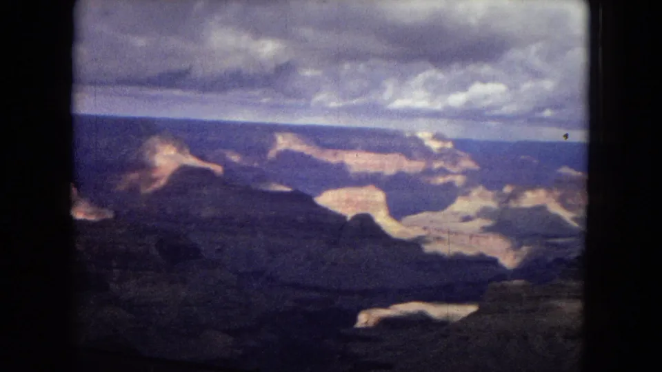 1967 8mm frame showing a broad Grand Canyon panorama with dark rock formations, distant walls, cloud cover, and a pale reflective area near the lower right.