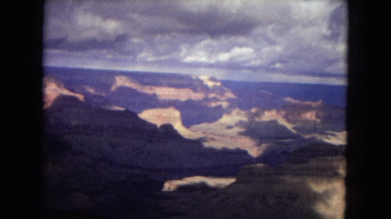 1967 8mm home-movie view of the Grand Canyon, with layered canyon walls, distant sunlit rock, dark foreground ridges, and a heavy cloud ceiling inside a projector-framed image.