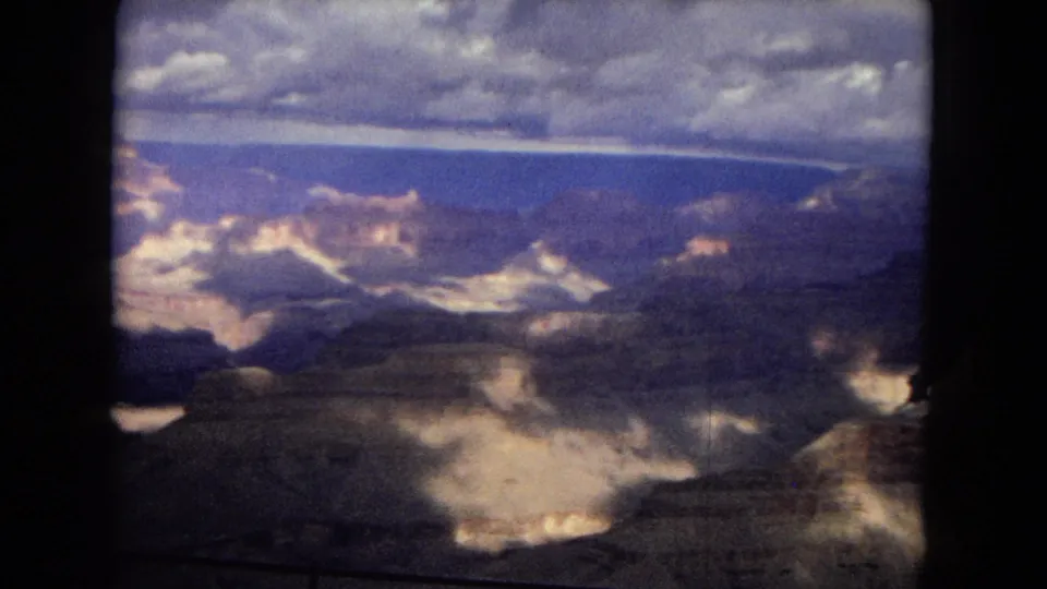 1967 8mm view across the Grand Canyon, with distant mesas, layered rock faces, dark canyon shadows, and a mottled sky softened by film grain and color shift.