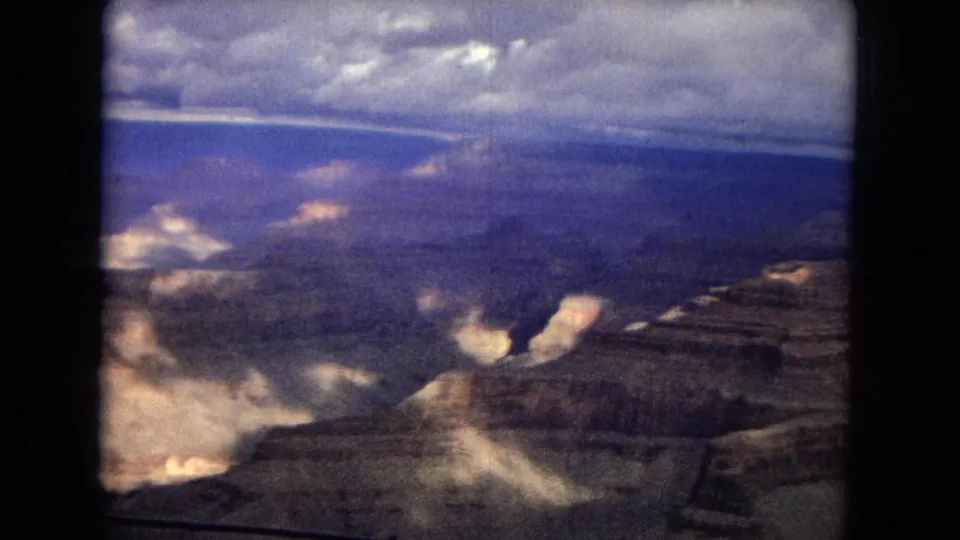 1967 home-movie frame looking over the Grand Canyon, where broad shadowed terrain, pale rock highlights, a blue-gray horizon, and bright cloud patches appear within the projected film border.