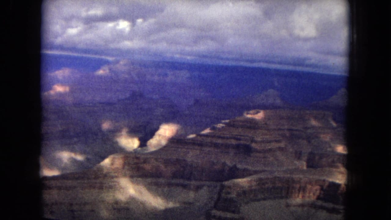 1967 8mm aerial view over the Grand Canyon, with layered rock formations, cloud shadows, and soft film grain visible across the frame.