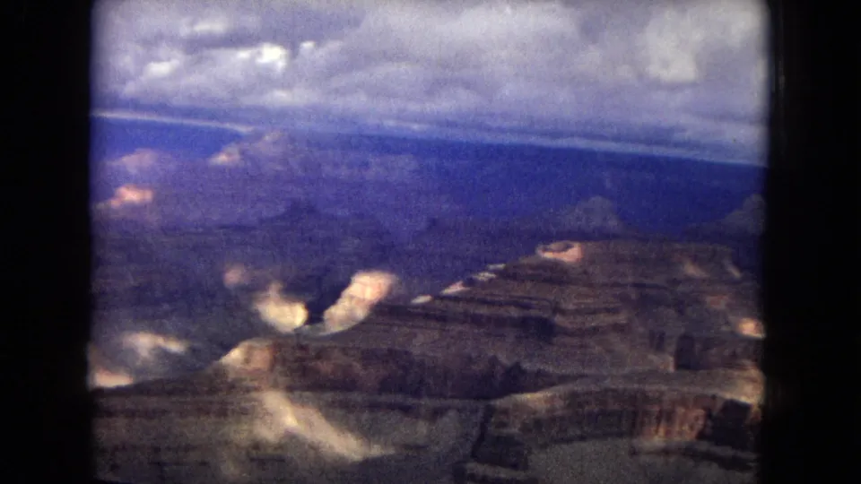 1967 8mm aerial view over the Grand Canyon, with layered rock formations, cloud shadows, and soft film grain visible across the frame.
