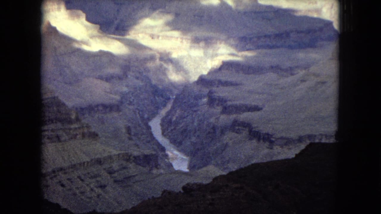 1967 8mm view of the Grand Canyon with the Colorado River winding through deep, layered rock formations under a bright clouded sky.