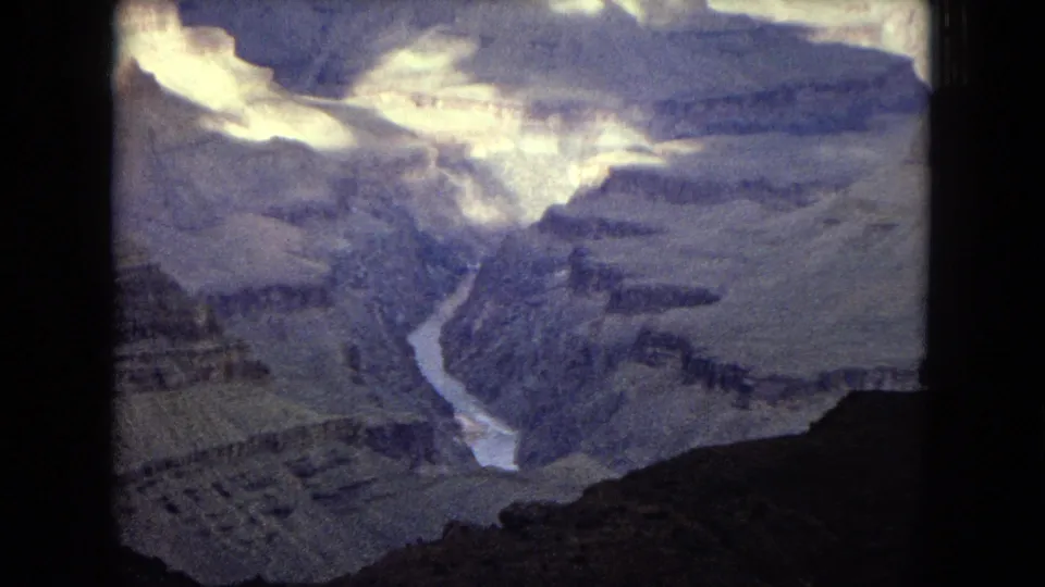 1967 8mm view of the Grand Canyon with the Colorado River winding through deep, layered rock formations under a bright clouded sky.