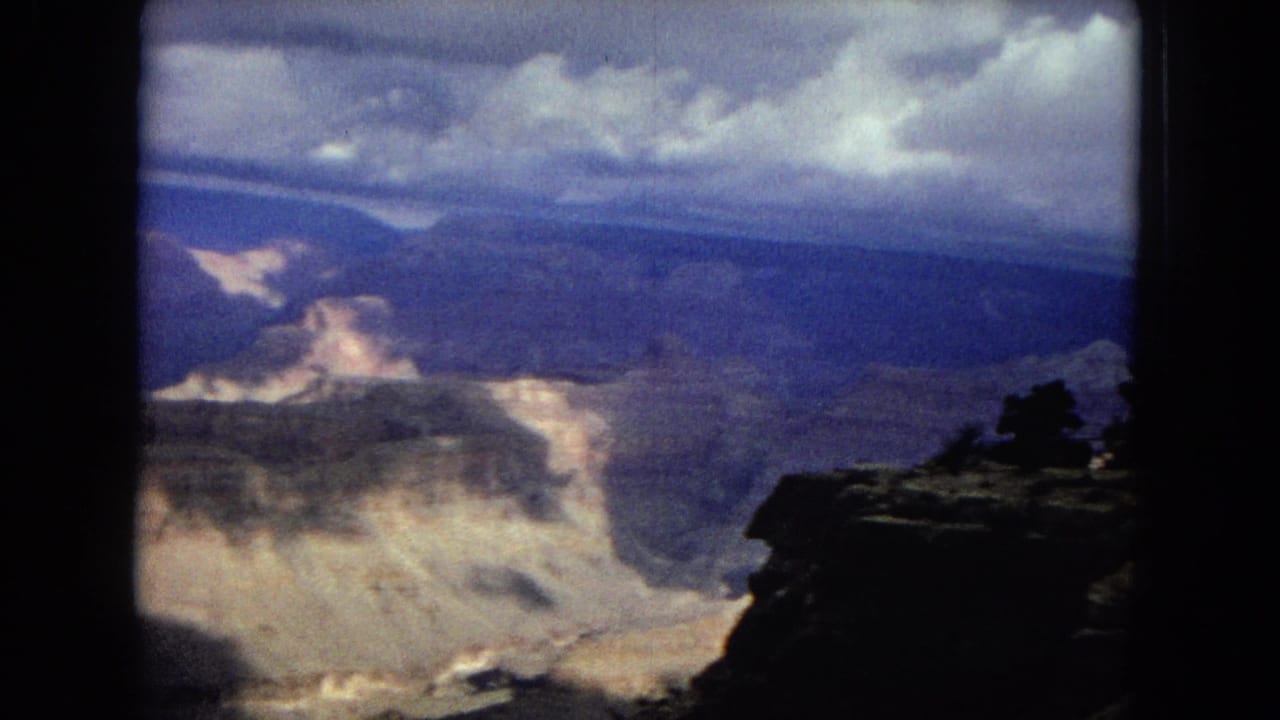 1967 8mm home-movie frame of the Grand Canyon with layered rock walls, a dark foreground ledge, and clouds casting uneven light across the canyon.