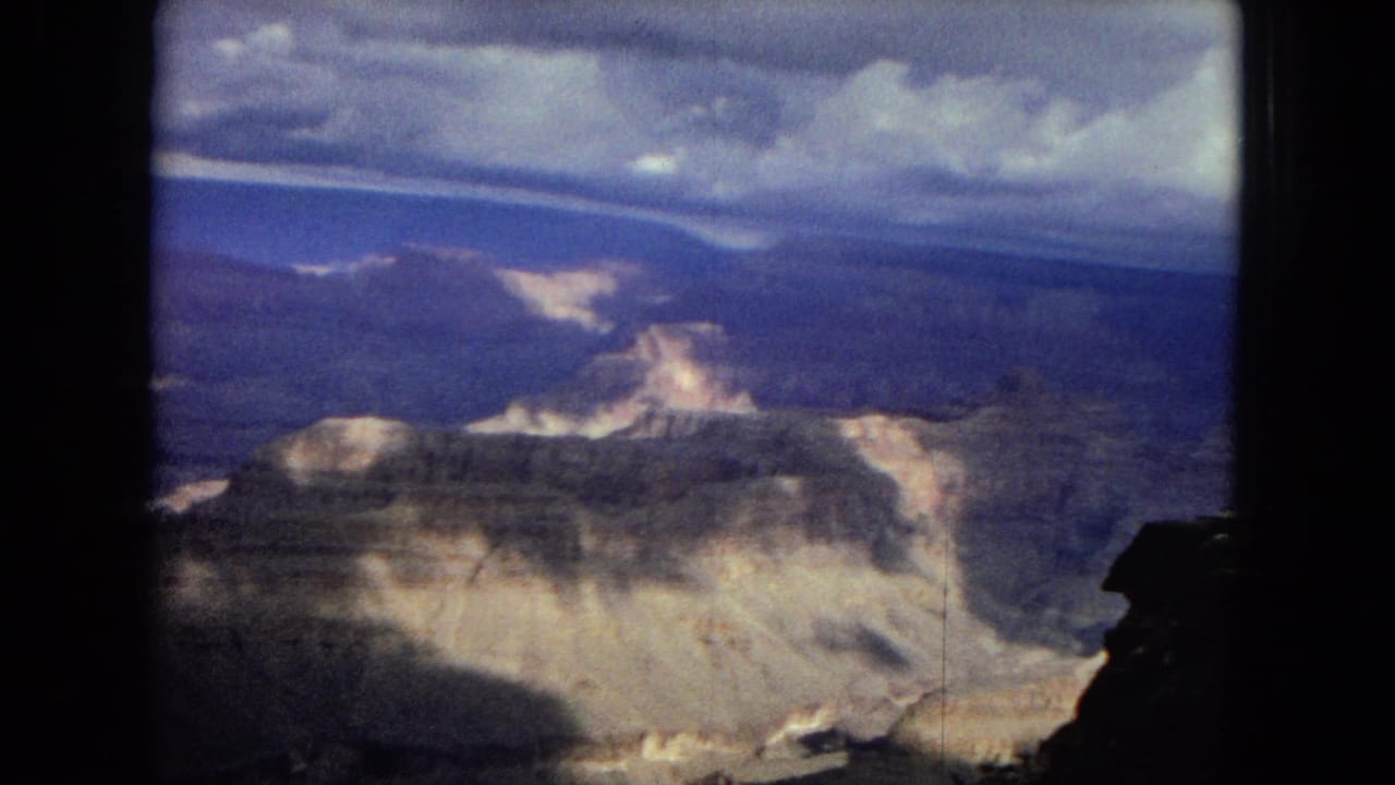 1967 8mm frame showing a wide Grand Canyon vista with pale eroded rock, distant blue-toned ridges, and dramatic cloud cover above.