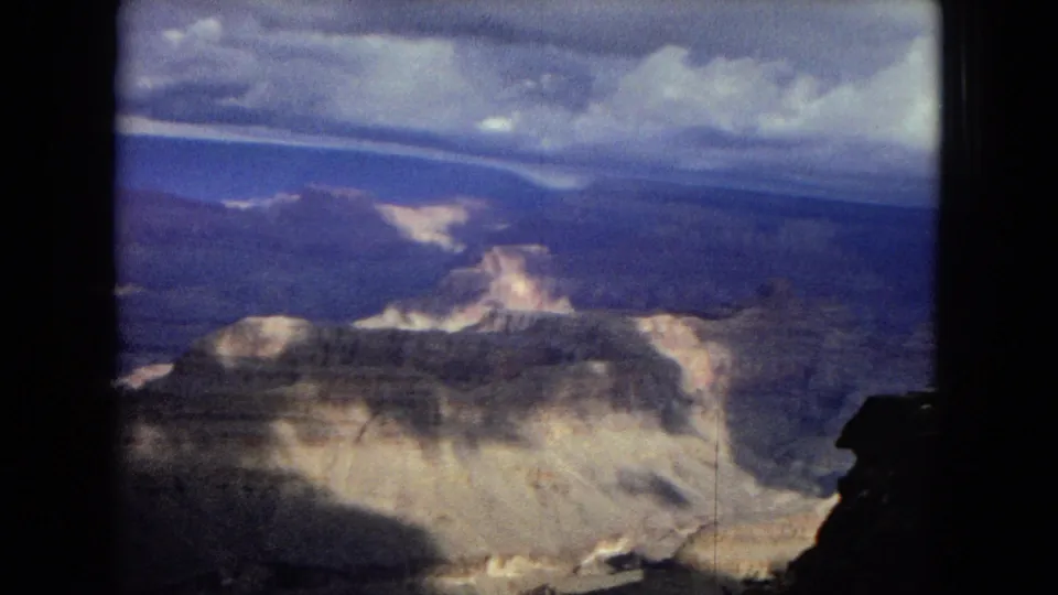 1967 8mm frame showing a wide Grand Canyon vista with pale eroded rock, distant blue-toned ridges, and dramatic cloud cover above.