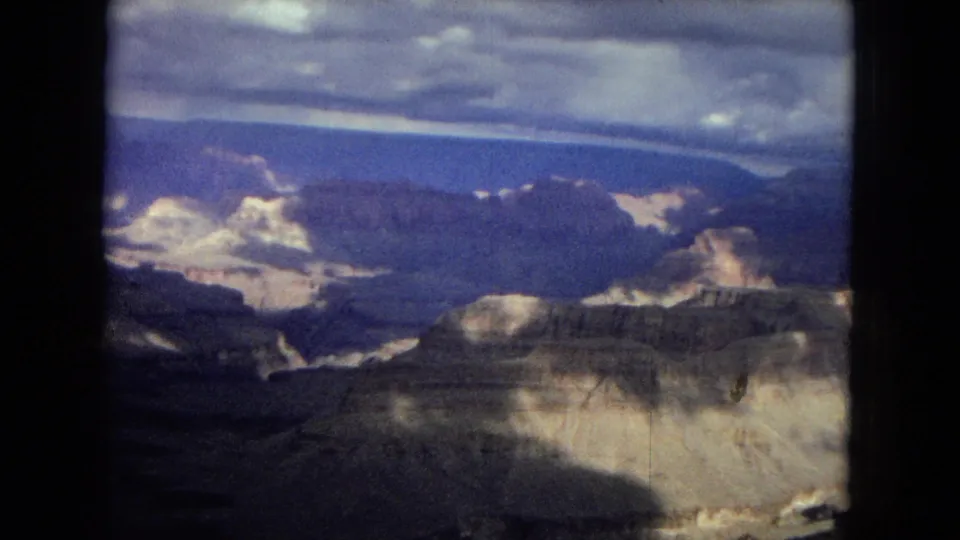 1967 8mm home-movie frame of the Grand Canyon panorama, with distant cliffs, bright exposed rock faces, and a broad cloudy sky across the horizon.