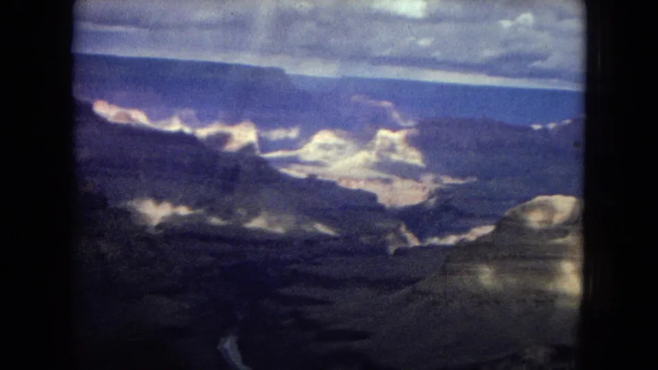 1967 8mm frame showing a darker Grand Canyon view with sunlit ridges, deep shadowed ravines, and a cloudy horizon in the distance.