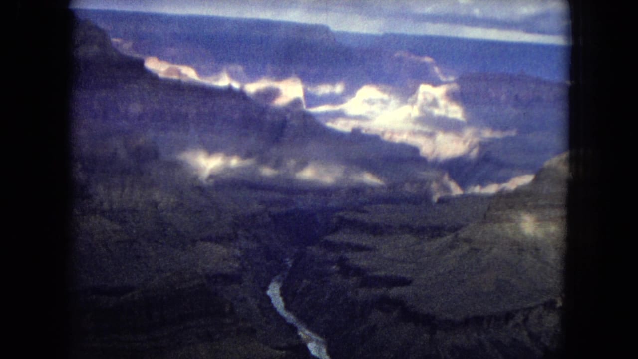 1967 8mm aerial view over the Grand Canyon, showing layered canyon walls, a bright winding river, and a clouded sky through dark film-frame edges.