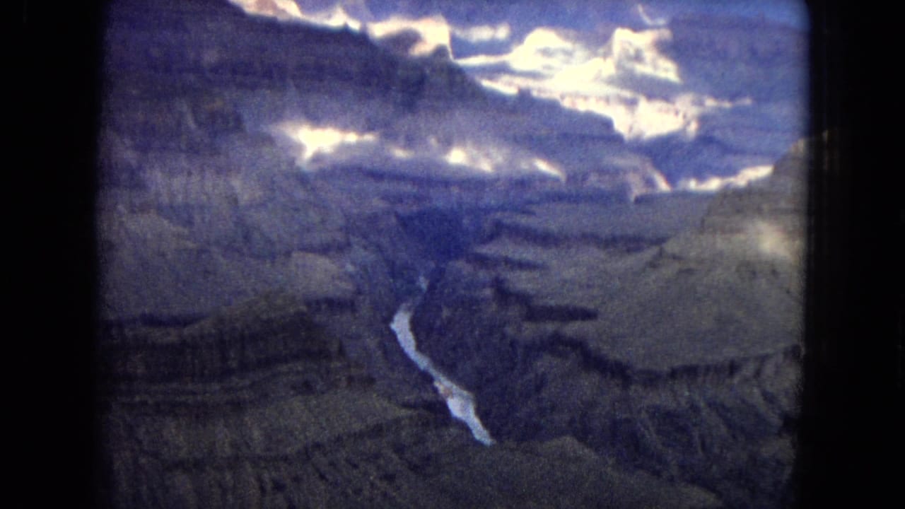 Aerial Grand Canyon view from 1967 home-movie footage, with broad rock terraces, a narrow river far below, and bright cloud glare near the top of frame.