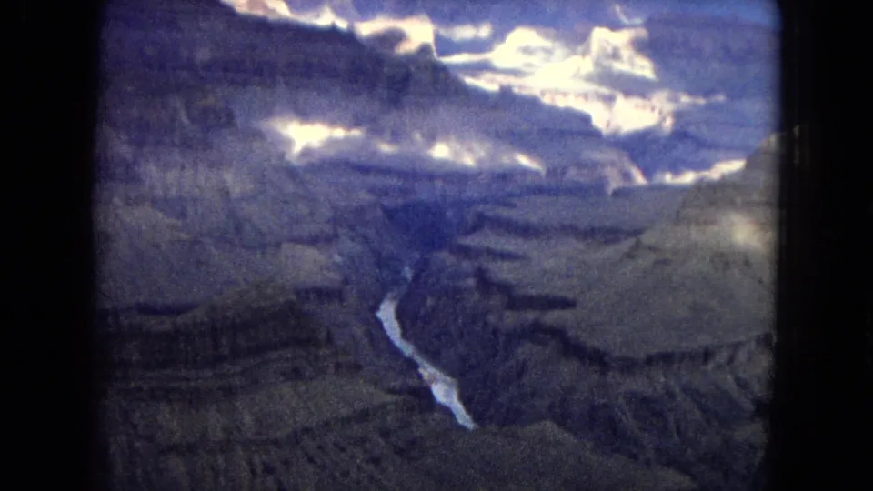 Aerial Grand Canyon view from 1967 home-movie footage, with broad rock terraces, a narrow river far below, and bright cloud glare near the top of frame.