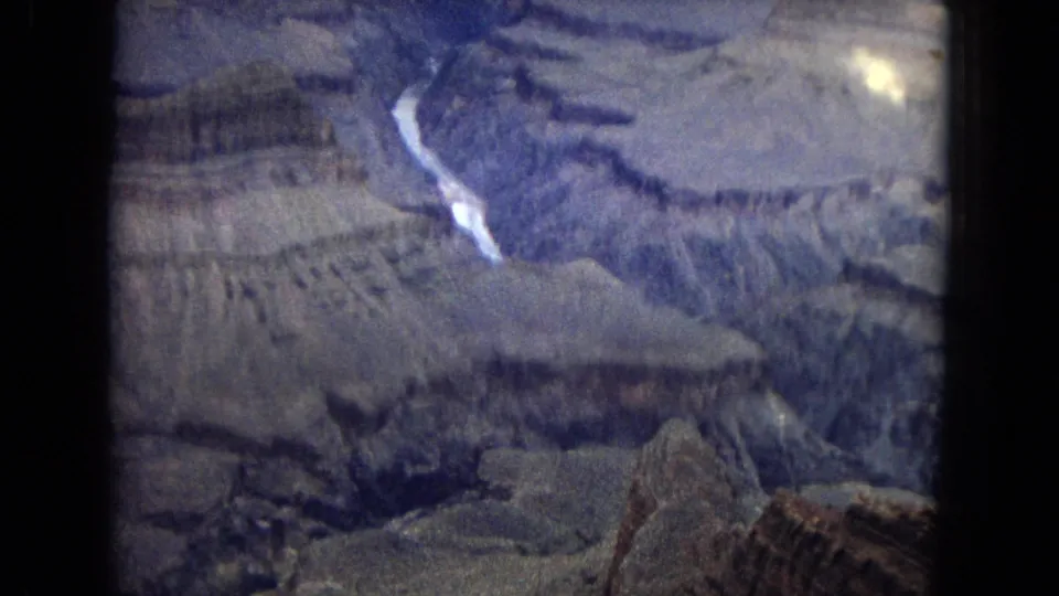 1967 8mm aerial footage looking down into the Grand Canyon, where layered ridges and a bright river curve through a hazy, grainy frame.