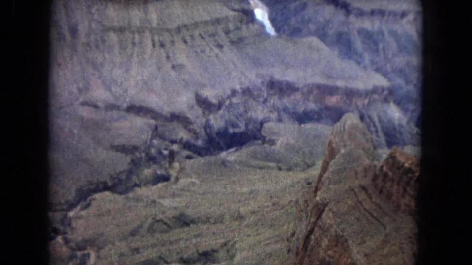 Aerial 1967 Grand Canyon home-movie frame showing rocky formations, broad ledges, a visible river segment near the top, and heavy 8mm gate-edge darkness.