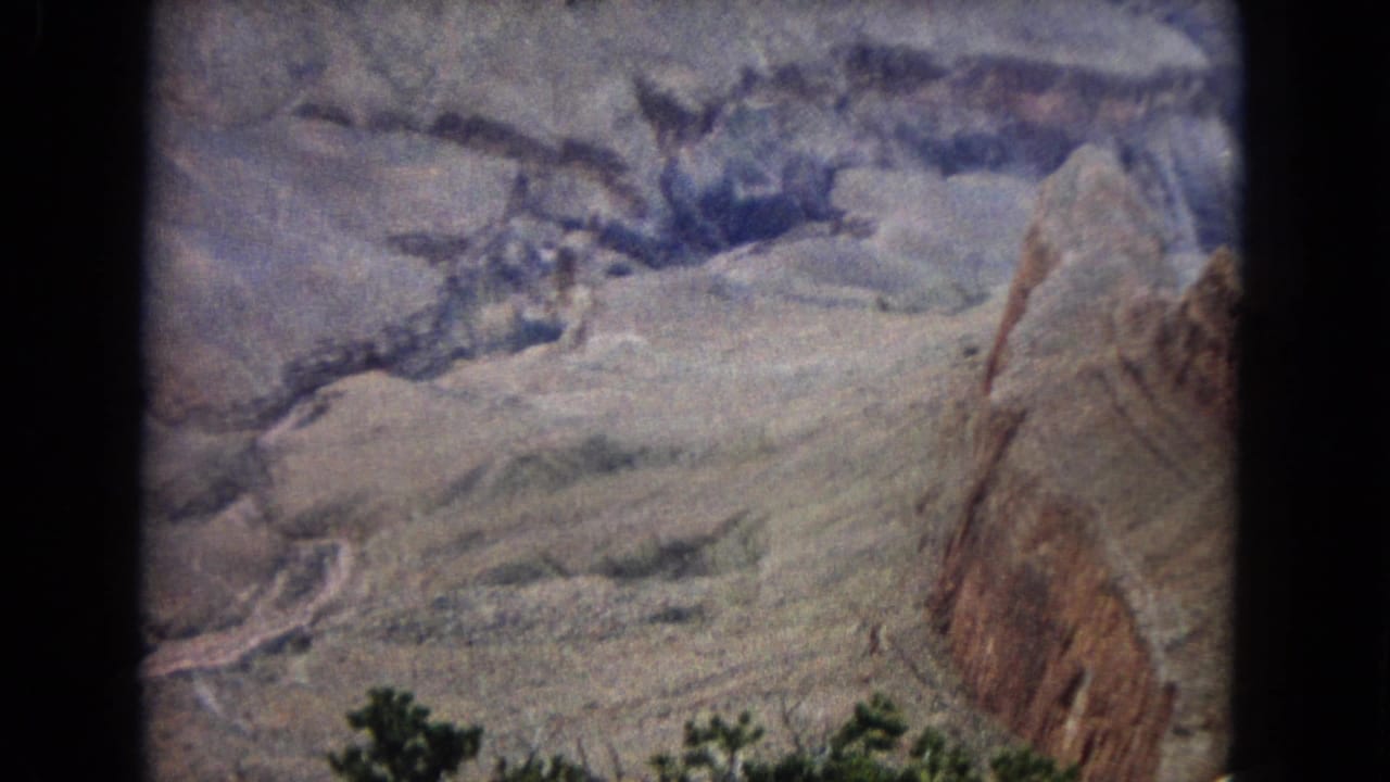 1967 8mm home-movie frame of a desert canyon in the United States, showing pale layered rock, a winding river or wash below, and dark green shrubs in the foreground.
