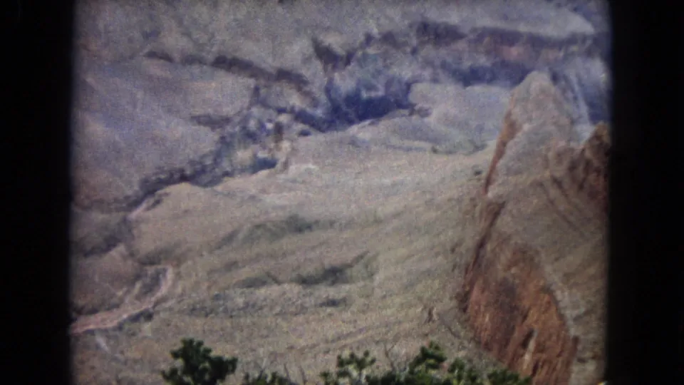 1967 8mm home-movie frame of a desert canyon in the United States, showing pale layered rock, a winding river or wash below, and dark green shrubs in the foreground.