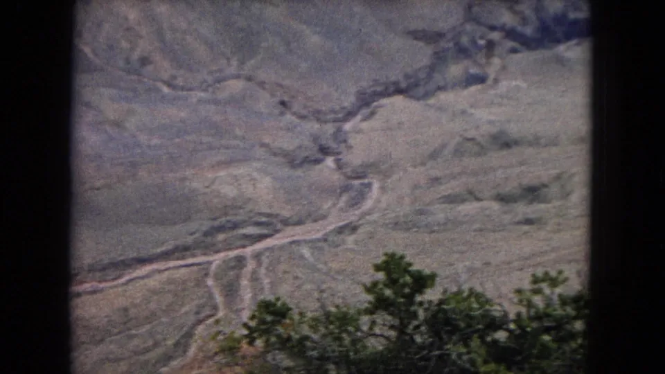1967 8mm home-movie frame of a broad desert canyon view, with a branching river or wash cutting through pale rock layers and sparse shrubs in the lower foreground.