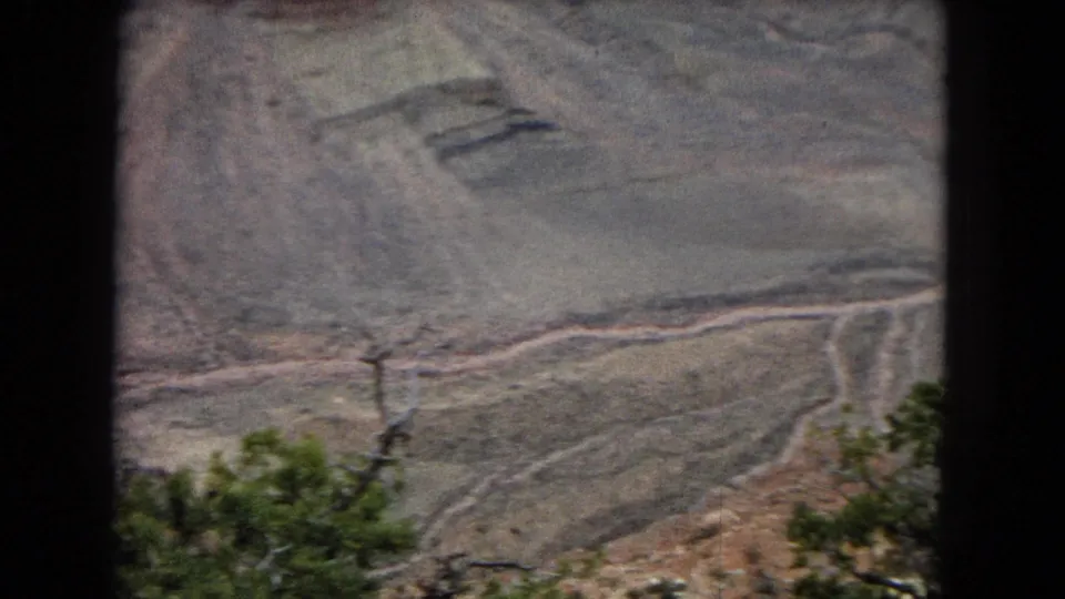 1967 8mm home-movie frame showing a muted desert canyon slope, a pale winding channel or track below, and dark green shrubs partly obscuring the lower frame.