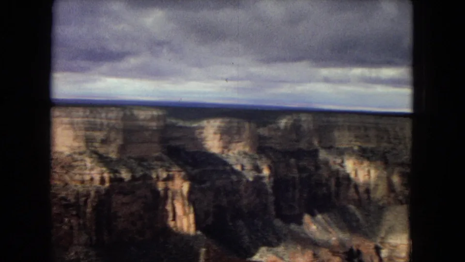 1967 8mm home-movie view across layered Grand Canyon formations, with a gray sky, distant rim, and dark shadowed canyon walls.