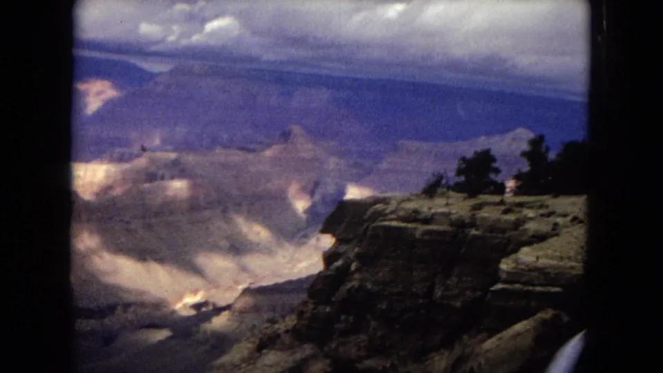 1967 8mm home-movie frame from a Grand Canyon overlook, showing a rocky ledge with small trees, distant canyon ridges, and patches of sun under cloud cover.