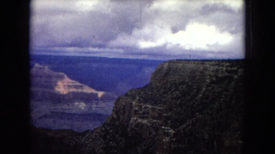1967 Arizona 8mm frame showing a rocky canyon wall at right, layered canyon terrain in the distance, and a cloudy sky within rounded film borders.