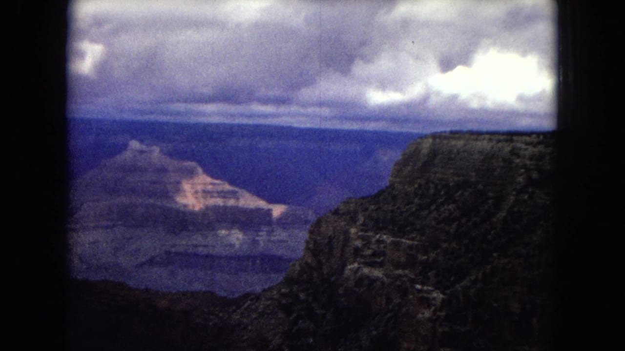 1967 8mm home-movie frame of the Grand Canyon from a high overlook, with layered canyon formations, dark foreground rock, and heavy clouds above.