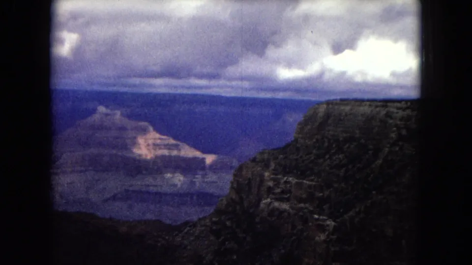 1967 8mm home-movie frame of the Grand Canyon from a high overlook, with layered canyon formations, dark foreground rock, and heavy clouds above.