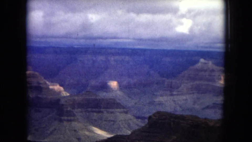 High overlook view of the Grand Canyon in 1967 8mm footage, with layered ridges receding into blue haze and clouds stretching across the horizon.