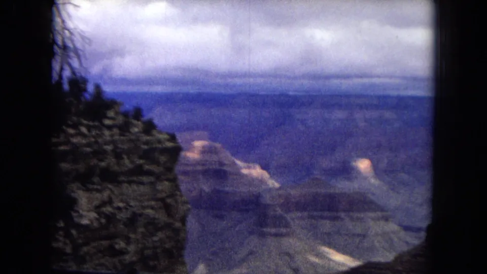 1967 8mm frame of the Grand Canyon with a dark foreground rock face at left, distant formations, muted blue canyon walls, and a cloudy sky.