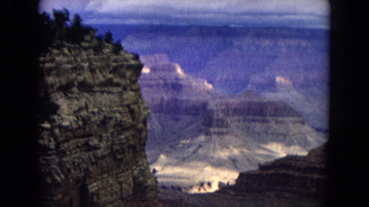 1967 8mm home-movie view of the Grand Canyon, with a dark rock edge framing layered cliffs, distant mesas, and a cloudy blue sky.