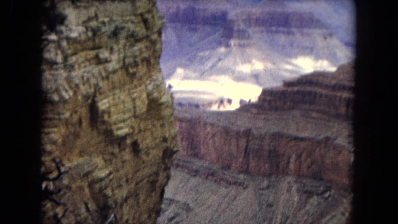 1967 8mm view from a Grand Canyon overlook, showing a tall textured rock face in the foreground and layered canyon walls receding into bright clouded light.