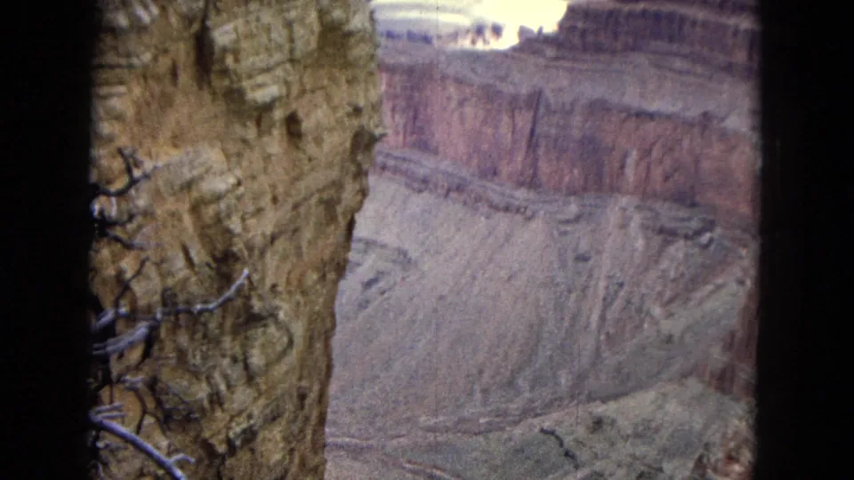 1967 8mm home-movie frame of the Grand Canyon, with textured foreground rock and dry branches looking across a pale canyon basin and reddish cliff band.
