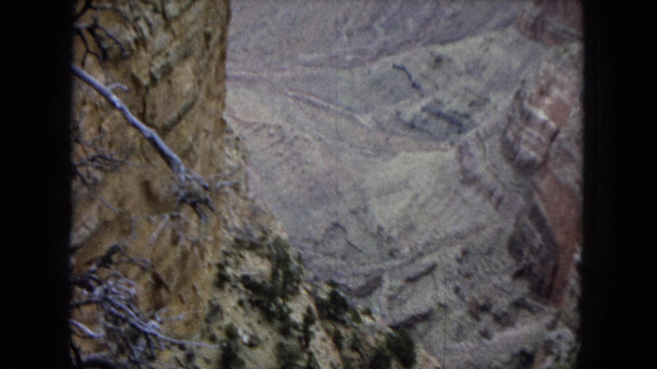 1967 Arizona 8mm frame showing a canyon overlook view with layered rock, pale eroded slopes, dry brush, and dark gate edges from the film scan.